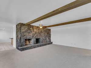 Unfurnished living room featuring carpet, tile patterned floors, ceiling fan, beamed ceiling, and a stone fireplace