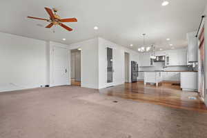 Unfurnished living room with ceiling fan with notable chandelier, a sink, baseboards, light carpet, and recessed lighting