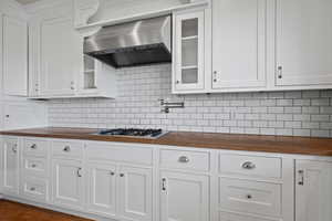 Kitchen with glass insert cabinets, butcher block counters, white cabinetry, gas cooktop, and wall chimney exhaust hood