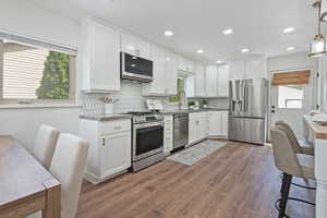 Kitchen with stainless steel appliances, a large single bowl Blanco sink, wood finished floors, white cabinetry, and recessed lighting