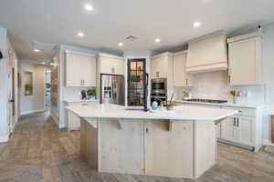 Kitchen featuring appliances with stainless steel finishes, custom range hood, a kitchen island with sink, light countertops, and backsplash