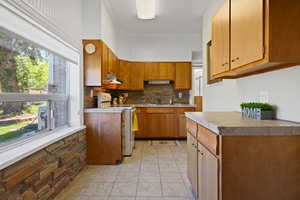 Kitchen with electric range, under cabinet range hood, tasteful backsplash, light tile patterned flooring, and brown cabinetry