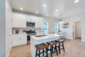 Kitchen with appliances with stainless steel finishes, a sink, light LVP flooring, white cabinetry, and a center island with sink