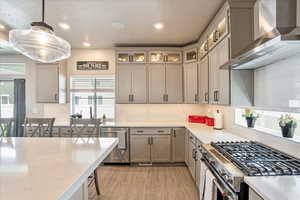 Kitchen featuring wall chimney exhaust hood, appliances with stainless steel finishes, light wood-style flooring, hanging light fixtures, and backsplash