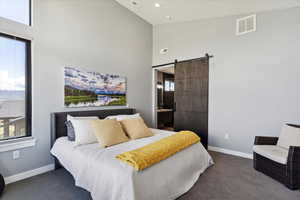 Carpeted bedroom featuring high vaulted ceiling, a barn door, baseboards, multiple windows, and ensuite bathroom
