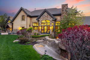 Back of house at dusk featuring a lawn, a chimney, french doors, stucco siding, and stone siding