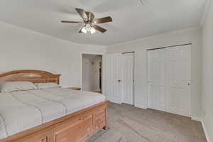 Bedroom with two closets, ornamental molding, light colored carpet, baseboards, and a ceiling fan