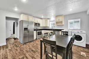 Kitchen with appliances with stainless steel finishes, washer / dryer, cream cabinetry, dark wood-type flooring, and a textured ceiling