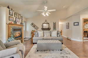 Living room with wood finished floors, a tiled fireplace, baseboards, a ceiling fan, and vaulted ceiling