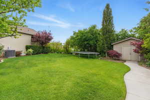 View of grassy yard with a trampoline, central AC unit, a storage shed, and an outbuilding