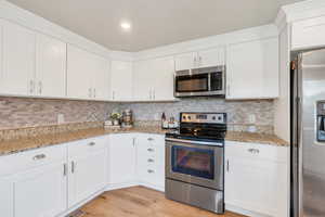 Kitchen featuring appliances with stainless steel finishes, white cabinetry, tasteful backsplash, light wood finished floors, and light stone counters
