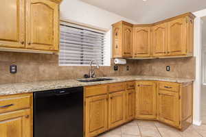 Kitchen featuring black dishwasher, a sink, tasteful backsplash, light tile patterned flooring, and light stone counters