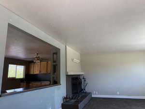 Unfurnished living room with a textured ceiling, baseboards, dark colored carpet, a ceiling fan, and a fireplace with raised hearth