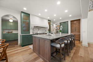 Kitchen with green cabinetry, light wood-style floors, tasteful backsplash, an island with sink, and light stone counters