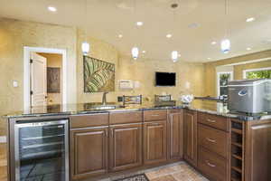 Kitchen featuring wine cooler, a sink, stone tile floors, a textured wall, and dark stone counters