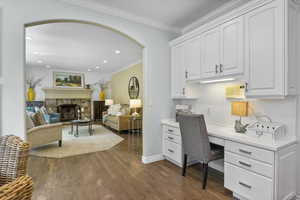 Office with arched walkways, built in desk, dark wood-type flooring, crown molding, and a stone fireplace