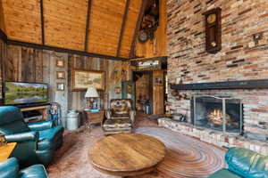 Carpeted living area with high vaulted ceiling, wooden walls, beam ceiling, a brick fireplace, and wood ceiling