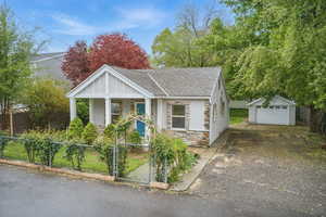 Bungalow-style house featuring covered porch, a fenced front yard, stone siding, roof with shingles, and a detached garage