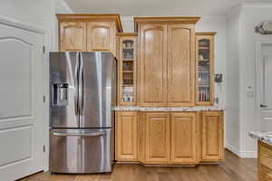 Kitchen featuring stainless steel refrigerator with ice dispenser, ornamental molding, light stone countertops, dark wood-type flooring, and glass insert cabinets