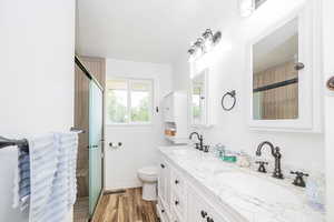 Full bath with double vanity, toilet, wood finished floors, a shower with shower door, and baseboards