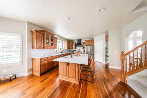 Kitchen featuring custom range hood, freestanding refrigerator, brown cabinetry, light countertops, and wood finished floors