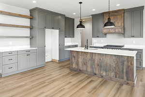 Kitchen with open shelves, light wood finished floors, recessed lighting, a center island with sink, and hanging light fixtures