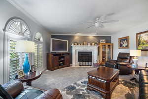 Carpeted living area with a ceiling fan, crown molding, a fireplace with flush hearth, and baseboards