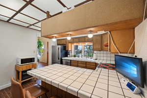 Kitchen featuring stainless steel appliances, tile counters, baseboards, brown cabinetry, and wallpapered walls