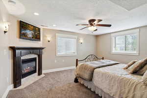 Bedroom with a textured ceiling, carpet flooring, a fireplace with flush hearth, baseboards, and a ceiling fan