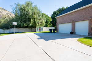 View of side of property featuring driveway, a garage, brick siding, fence, and a shingled roof