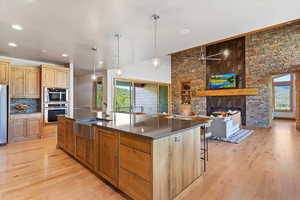 Kitchen with dark countertops, light wood-type flooring, a stone fireplace, an island with sink, and a sink