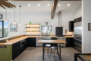 Kitchen with built in appliances, butcher block counters, wall chimney exhaust hood, dark cabinets, and beamed ceiling