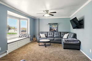 Living room featuring a baseboard heating unit, light carpet, crown molding, ceiling fan, and baseboards