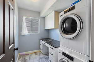 Laundry room with stacked washer and clothes dryer, cabinet space, light marble finish flooring, and baseboards