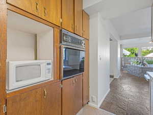 Kitchen featuring white microwave, wall oven, baseboards, brown cabinets, and stone finish flooring