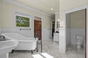 Full bathroom featuring a wainscoted wall, marble finish flooring, crown molding, a soaking tub, and toilet