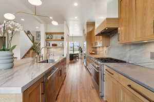 Kitchen with high quality appliances, a sink, wall chimney range hood, open shelves, and light stone counters