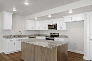 Kitchen with stainless steel appliances, light stone counters, a center island, white cabinets, and dark wood-type flooring