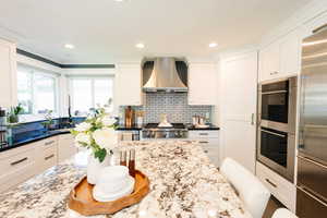 Kitchen featuring wall chimney range hood, white cabinetry, stainless steel appliances, light stone counters, and decorative backsplash