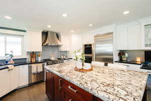 Kitchen featuring wall chimney range hood, appliances with stainless steel finishes, dark wood-type flooring, white cabinetry, and glass insert cabinets