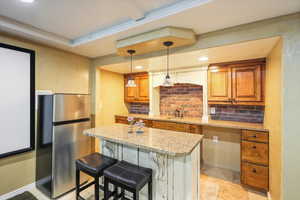 Kitchen featuring freestanding refrigerator, a sink, light stone countertops, brown cabinetry, and a breakfast bar