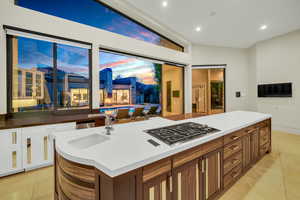 Kitchen with light tile patterned floors, stainless steel gas cooktop, a center island with sink, brown cabinets, and recessed lighting