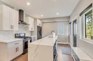 Kitchen with appliances with stainless steel finishes, wall chimney range hood, a sink, decorative backsplash, and an island with sink
