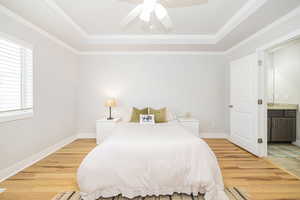 Bedroom with light wood-style flooring, a raised ceiling, connected bathroom, and crown molding
