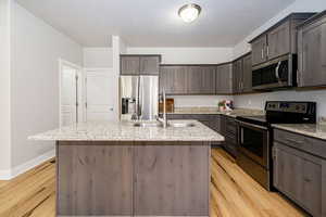 Kitchen featuring stainless steel appliances, a sink, light wood finished floors, dark brown cabinets, and an island with sink