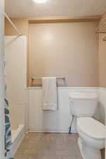 Bathroom featuring a textured ceiling, a wainscoted wall, toilet, stone finish floors, and shower / bath combo