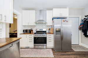 Kitchen with appliances with stainless steel finishes, butcher block counters, wall chimney exhaust hood, dark wood-type flooring, and tasteful backsplash