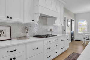 Kitchen with stainless steel gas stovetop, light wood-style flooring, decorative backsplash, white cabinets, and light stone counters