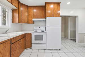 Kitchen with white appliances, brown cabinetry, light countertops, under cabinet range hood, and light tile patterned flooring