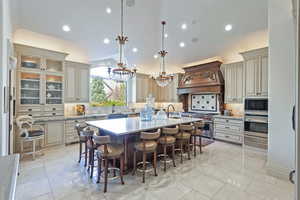Kitchen with appliances with stainless steel finishes, custom range hood, cream cabinetry, an island with sink, and vaulted ceiling